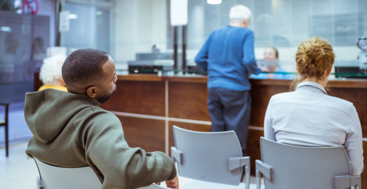 Rear view of people sitting in a hospital waiting room. One person is checking in at a main desk.