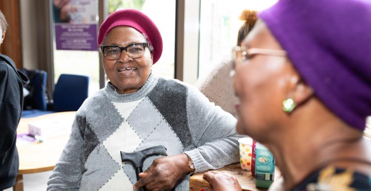 Two older black woman talking and smiling. Both are wearing berets and holding walking sticks.