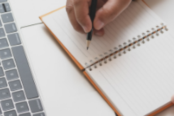 Close-up of a person's hand writing in a notepad, in front of a laptop.