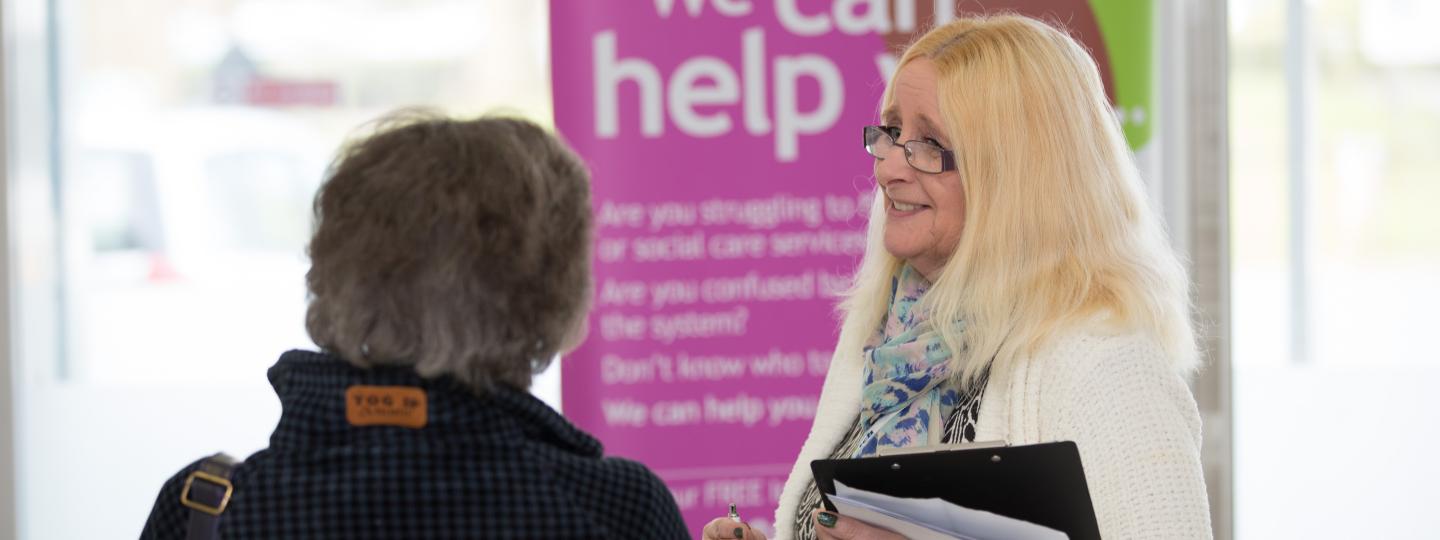 Woman standing in front of a banner that says 'we can help' speaking to a member of the public